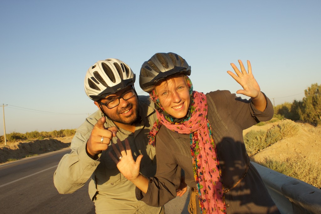 Annika und Roberto ohne Fahrräder aber mit Helmen auf, auf einer Straße die von trockener Landschaft umgeben ist