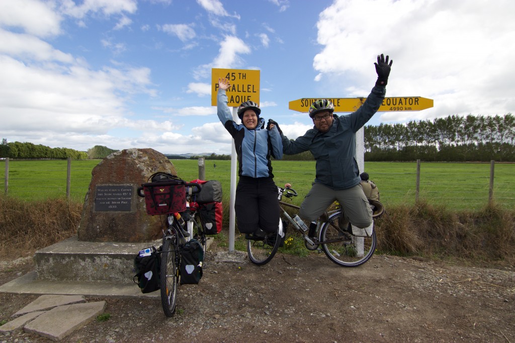 Annika and Roberto jumping in the air. In the background two street signs. Left: "45th Parallel Plaque" and right two arrows. The one facing left shows the way to the south pole and the other one to the equator.